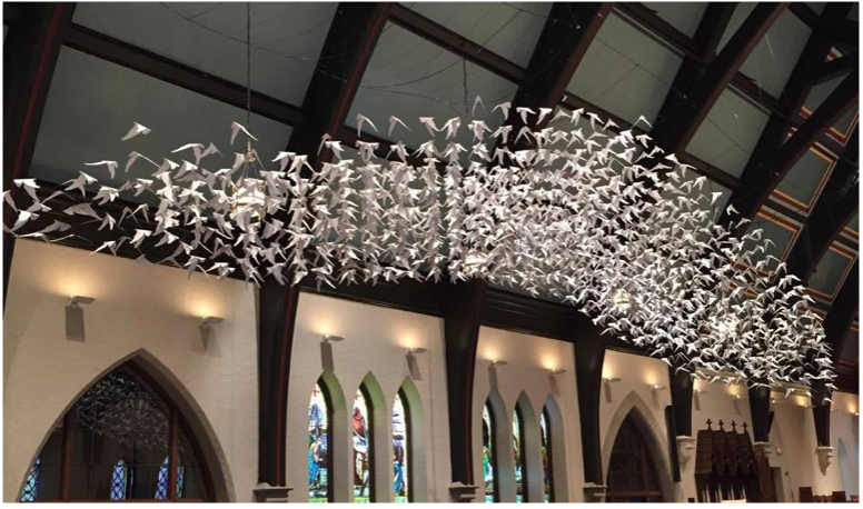 "Countless" origami doves positioned over the pews in a church nave, as if alighting on those sitting there.